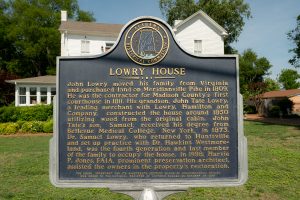 A black historical marker depicts the written history of the Lowry House in gold type. The house is seen just behind the marker.