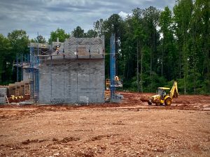 Crews build a concrete-block wall at the site of the future Huntsville Amphitheater at MidCity District. There is a yellow backhoe at the right and trees in the background.