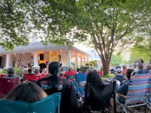 A few dozen people sit in lawn chairs on the front lawn of a house in the Five Points area. They are looking toward a front porch, where music artists are performing. They are under a large tree.