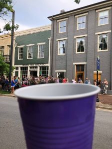 A purple plastic cup is seen in the foreground and there is a group of people far away in front of a gray building.