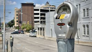 A parking meter is in the foreground on a city street with buildings in the background. There's a sticker on the meter advising how to pay by phone.