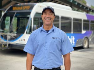 A portrait of Orbit bus operator Alvin Franklin. He's wearing a dark blue cap, a light-blue Huntsville Transit uniform top and he's standing in front of an Orbit bus at the Huntsville Transit station in Huntsville.