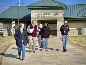 A man in tan pants and a red vests talks to an African-American woman and other people as they walk away from John Hunt Park volleyball complex