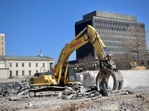 A piece of machinery picks up remnants of the municipal parking garage under a blue sky. The Madison County Courthouse can be seen in the background.