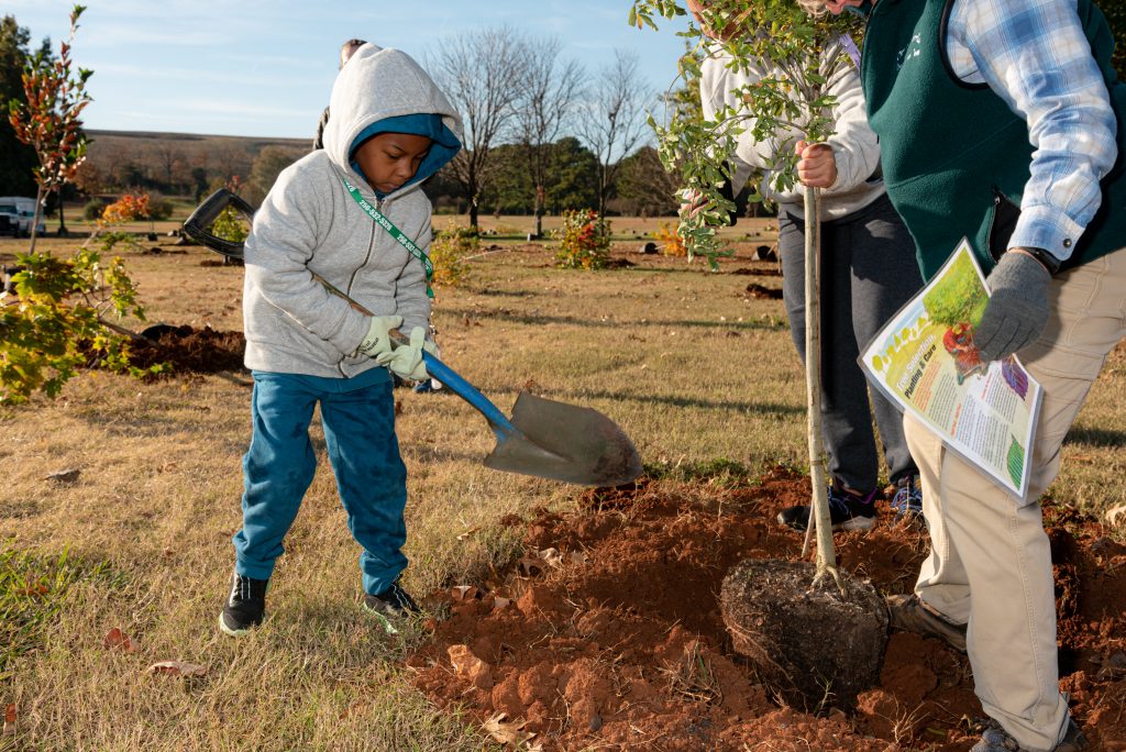 Child and his mother help a landscape management worker plant trees in John Hunt Park