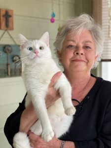 A woman holds up a white, cross-eyed cat. The woman is wearing a black shirt and has gray hair.