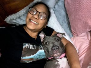 A woman in a black T-shirt lays with a large gray and white dog. There are pillows in the background.