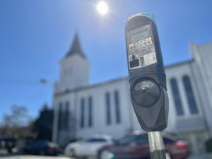 A parking meter in downtown Huntsville is seen with a church in the background. The sun is overhead. The meter is black and has a digital display on the front.