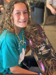 A young girl holds a signed skateboard. She has curly hair and is wearing a green shirt. The skateboard is multicolored and has Tony Hawk's signature in the middle.