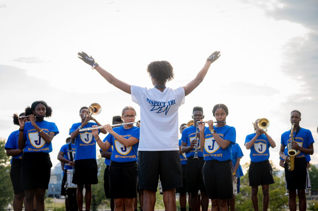 The drum major for the Mae C. Jemison High School Band lifts her arms to direct the band outside City Hall on Sept. 22, 2022. She is wearing a white T-shirt that says "Respect the Den." There are musicians in the background with instruments raised.  They are wearing blue T-shirts.