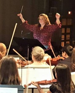 A woman lifts her arms to direct a group of young students playing violin.