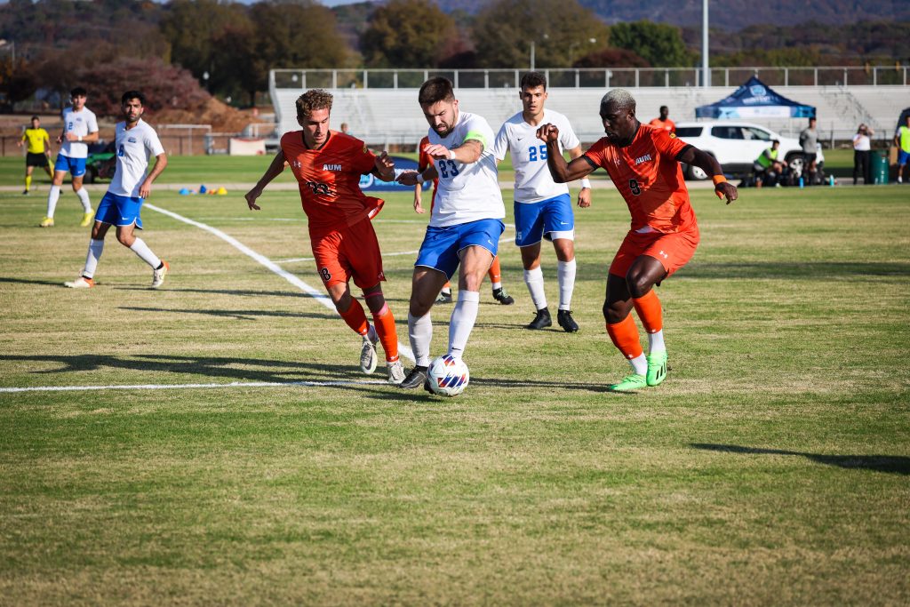 Four players play a soccer match on a field in Huntsville. Two players are in orange, while the other two are in white. They are trying to get possession of the ball.