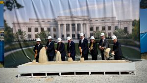 A group of people in dark suits and white hard hats throw shovels of sand during a groundbreaking event. Behind them is a rendering of the new federal courthouse.