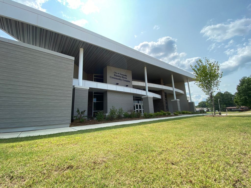 An exterior shot of the Sandra Moon Community Complex. The building is gray and white and very modern. There is a tree at right and green grass.