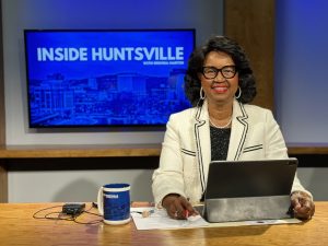 Brenda Martin, the Host of Inside Huntsville, poses at her anchor desk in front of a television screen.