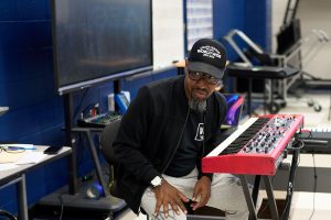 Kelvin Wooten sits behind a keyboard in a classroom at Lee High School. He is wearing a black jacket and black ballcap. His keyboard is red.