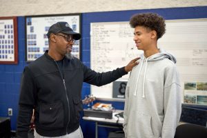 Multi-instrumentalist and Grammy award-winning producer Kelvin Wooten encourages a Lee High School student during a masterclass workshop. Wooten is wearing a black zip-up jacket and black ball cap. The student is wearing a gray sweatshirt.