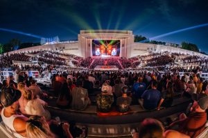 A shot of the Orion stage from back in the crowd. There are orange lights on stage and blue sky overhead. There is a crowd of people looking toward the stage.