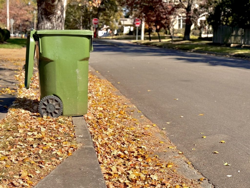 A green trash can sits on the corner of a curb surrounded by fallen leaves in downtown Huntsville.