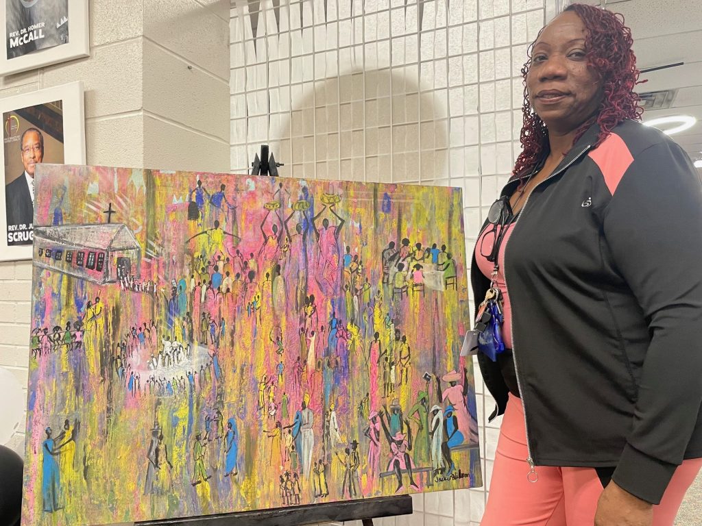 Jackie Wilson displays one of her abstract paintings, many of which celebrate Black history. She is wearing pink and a black jacket. The painting is very colorful.