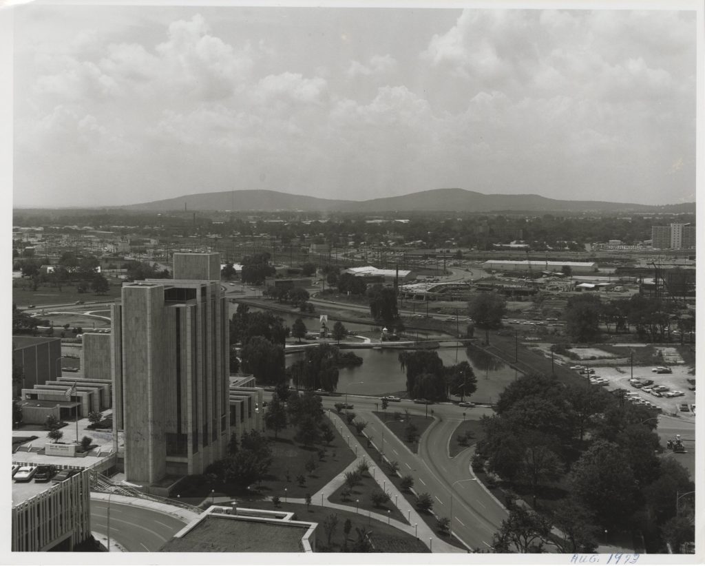 black and white aerial photo of city with tall building on the left, a road in the bottom center and a lake in the center with mountains in the background