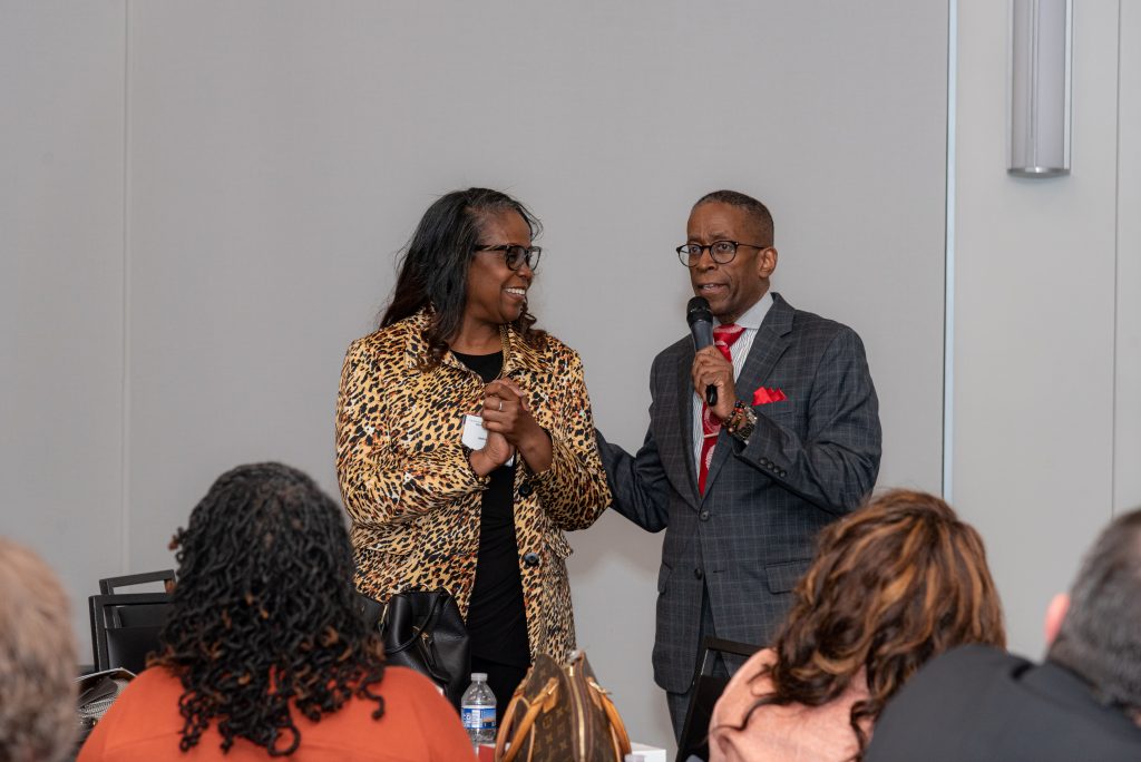 A man in a suit at right holds a microphone as he talks to a woman wearing an animal-print jacket, also in glasses. There are people in an audience watching them speak.