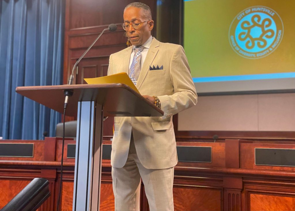 A man in a tan suit stands at a podium. Behind him are a dais and a big screen displaying the Huntsville Office of Diversity, Equity & Inclusion logo.
