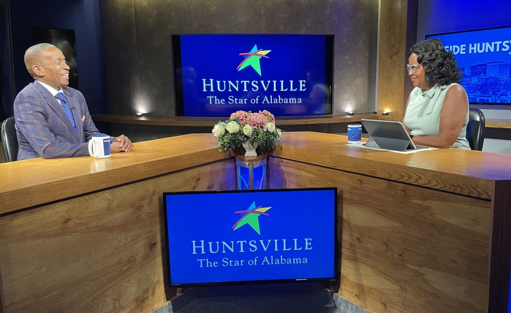 Two people sit at a desk looking at each other. There are TV screens all around, two of which have the City of Huntsville logo on it. The other says Inside Huntsville in all caps. The man is wearing a blue suit. The woman is wearing a light green dress.