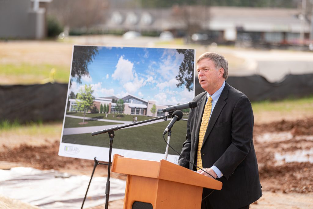 Man in blue coat and yellow tie speaks from wooden podium with building rendering in background