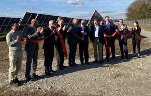 A group of people cut a red ribbon while standing on a gravel road. There are solar panels behind them and a chain fence.