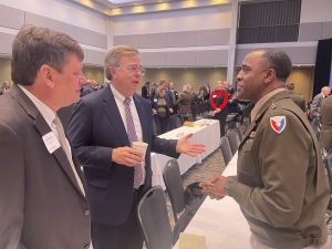two men in suites on left talking to Army general in uniform on right in large conference room