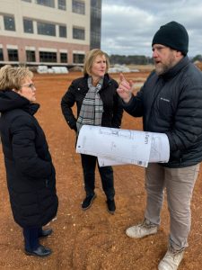 Two women at left stand in a field of dirt talking to a man at right holding architectural plans.