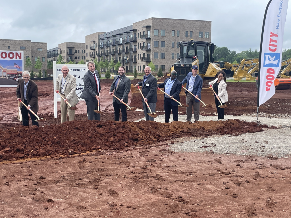 Group of people toss dirt at ceremonial groundbreaking ceremony with large building in background