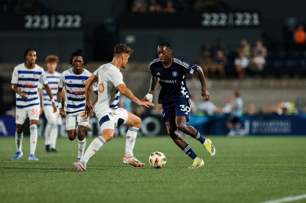 two players competing over ball at soccer game. player on right in blue uniform, player on left in white uniform
