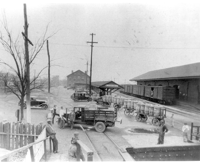 old black and white photo at the depot, from the 1930s, showing men standing around by the railroad tracks with their carts, model-t cars and a pickup truck , waiting to load barrels onto a train.
