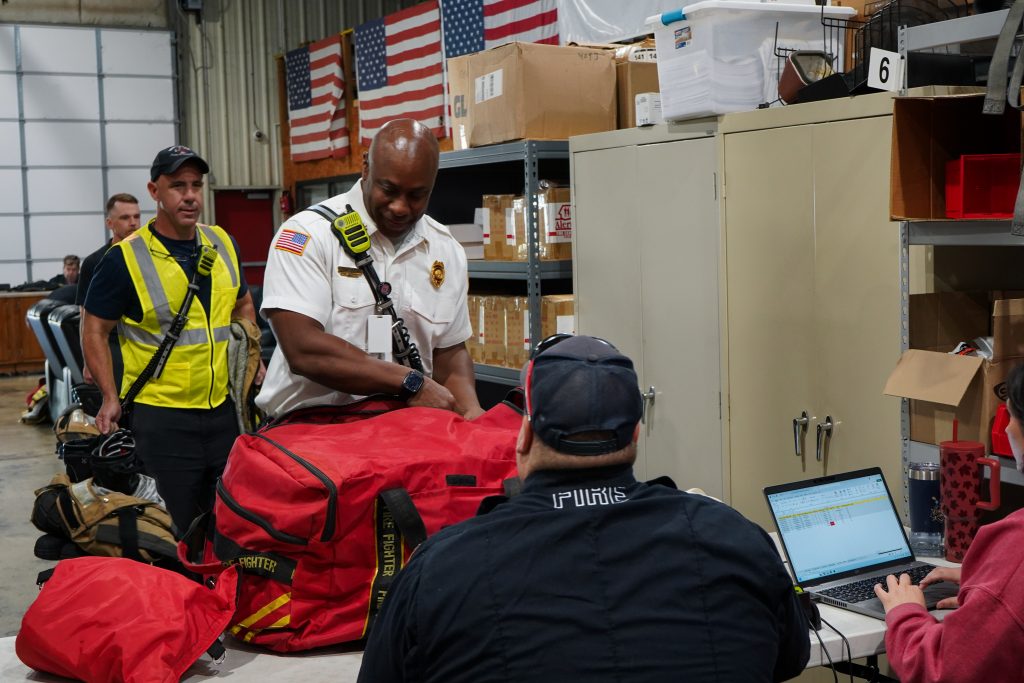 firefighter in white shirt presents red bag to be inspected by people seated at table