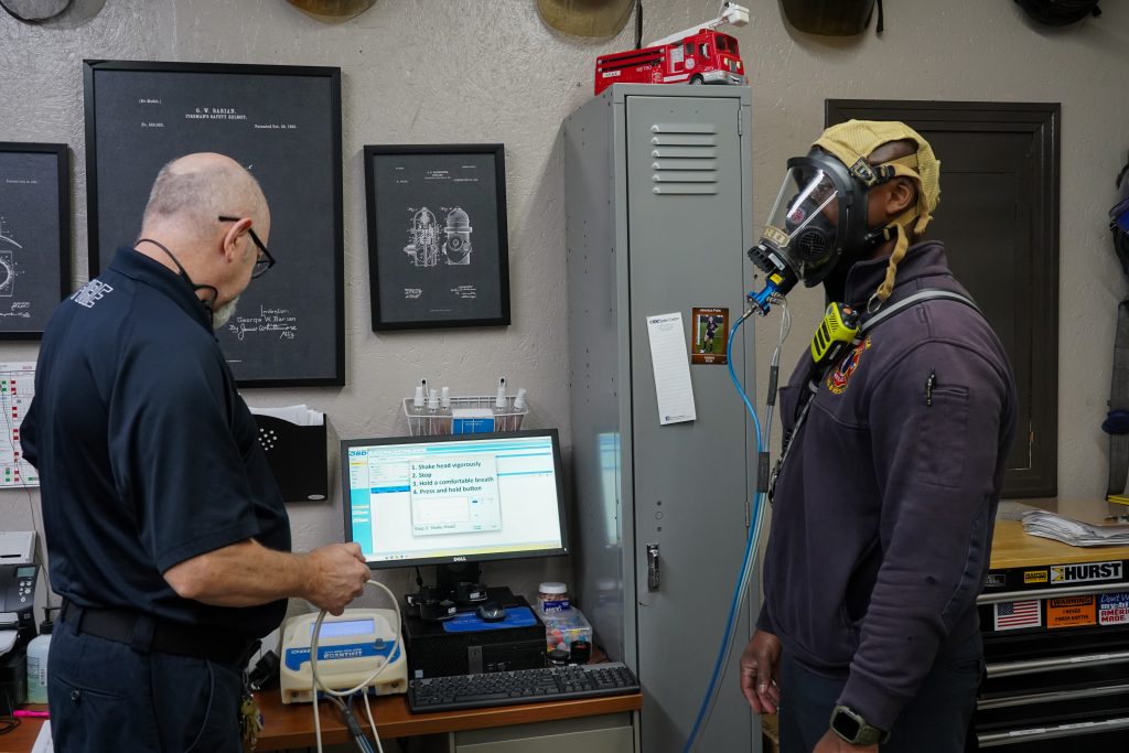 man on left looks at computer screen as man on right wears oxygen mask connected to white machine in middle
