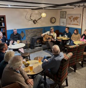 White male musician playing guitar to a small audience in a Scottish inn with deer antlers hanging on the wall
