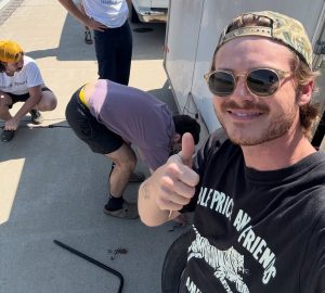 Young white male musician wearing sunglasses, standing with three other white male musicians as they repair a flat tire on their touring trailer